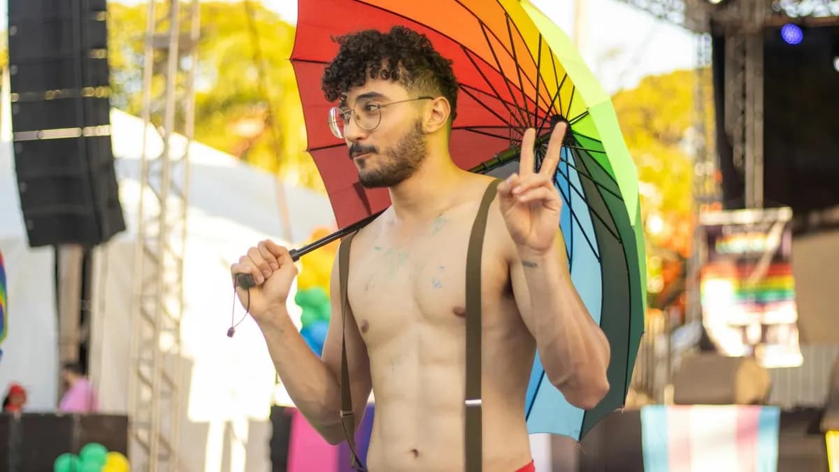 Shirtless man with a rainbow umbrella at a pride event, posing with a peace sign.