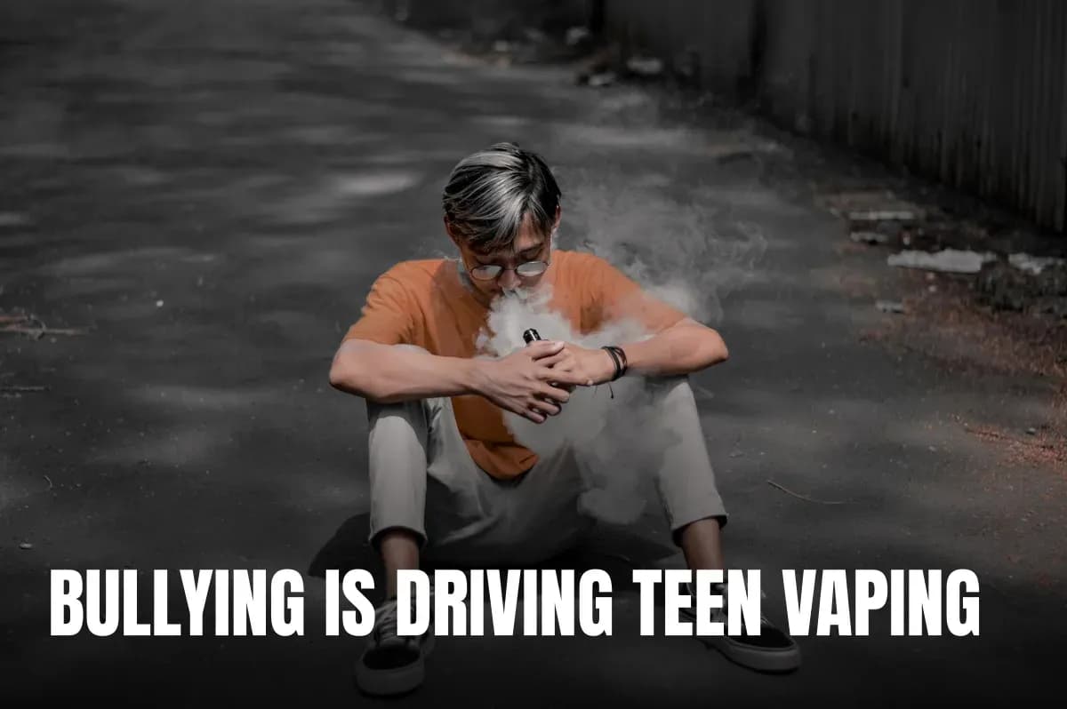 A young man wearing eyeglasses sits on an urban street while vaping, surrounded by smoke.